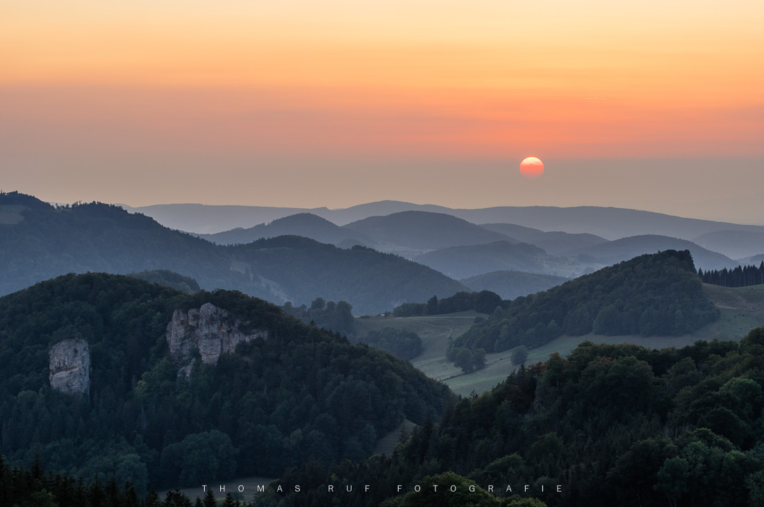 Dunstige Hügelsilhouetten im Baselland bei Sonnenuntergang – warme Lichtstimmung mit sichtbarer Sonne